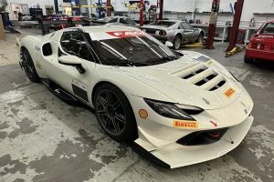 White Ferrari race car in a workshop garage with sponsor decals (Pirelli, Shell) on the nose and sides, and visible hood vents and aero details.
