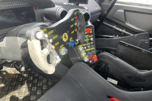 IMG_4767 – Sage Auto Sport Close-up of a Ferrari race car cockpit with a carbon-fiber steering wheel and labeled switches on the control panel, in a high-performance interior.