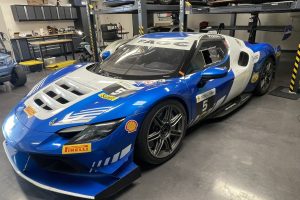 mikeM296 5 – Sage Auto Sport Blue and white Ferrari race car on a workshop floor, with a multi-level car lift and spare tires in the background.