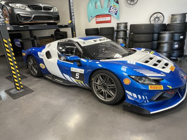 mikeM296 6 – Sage Auto Sport Blue and white race car with number 5 inside a garage full of stacked tires and a car on a lift above.