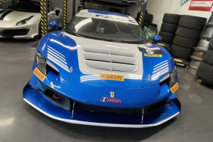mikeM296 7 – Sage Auto Sport Blue race car front in a garage, with Pirelli stickers and number 5, surrounded by stacked tires and a white car on a lift in the background.