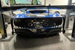 mikeM296 8 – Sage Auto Sport Blue race car on a hydraulic lift in a garage, with teal race car elevated above and spare tires nearby.