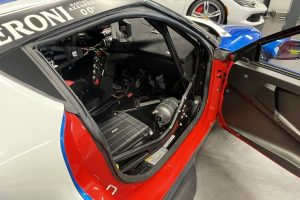 mikeM296 9 – Sage Auto Sport Red race car with the door open, exposing the cockpit wiring and interior components, with a white Ferrari in the background in a showroom.