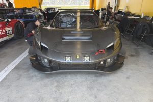 IMG_1572 - Copy – Sage Auto Sport Front view of a black Ferrari race car in a pit garage, technician adjusting the left front area beside other race cars and workstations in the background.