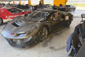 IMG_1573 – Sage Auto Sport Black carbon-fiber race car in a paddock under a canopy, with other race cars and tools in the background; a large rear wing and gold-rimmed wheels visible.