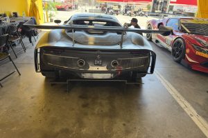 IMG_1575 – Sage Auto Sport Rear view of a matte black race car with a large rear wing, mesh grille, and dual exhausts in a pit garage; other race cars and tents visible in the background.