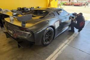 IMG_1576 – Sage Auto Sport Carbon-fiber race car in a yellow pit garage; mechanic crouches beside the cockpit working on the front fender. (informative)