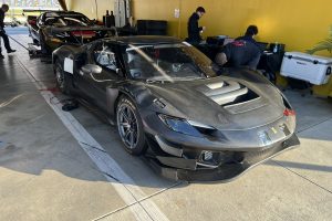 Black carbon-fiber Ferrari race car in a garage, with crew members working nearby and equipment in the background.