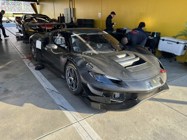 IMG_2282 – Sage Auto Sport Black carbon-fiber Ferrari race car in a garage, with crew members working nearby and equipment in the background.