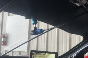 IMG_4467 – Sage Auto Sport Inside a car with a blue mounted device attached to the windshield and a monitor screen on the dashboard showing reflections of a showroom outside.