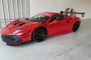 Red racing car in a garage with a large carbon fiber rear wing and black wheels, number 26 on the hood.