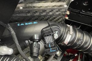IMG_4484 – Sage Auto Sport Close-up of a car engine bay showing a carbon-fiber panel, hoses, and electrical connectors, with a teal handwritten date on a black surface (8.11.2017-20).