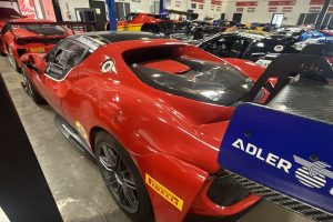 24 ferrari 296 301949-2 – Sage Auto Sport Indoor race shop with red Ferrari race cars parked linearly, showing sponsor decals and carbon-fiber rear sections.
